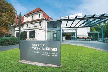 Auguste-Viktoria-Klinik Das Bild zeigt die Auguste-Viktoria-Klinik mit einem modernen Eingangsbereich und einem historischen Gebäude im Hintergrund. Im Vordergrund steht ein Schild mit dem Namen der Klinik, während eine Überdachung den Eingangsbereich überspannt.