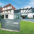 Auguste-Viktoria-Klinik Das Bild zeigt die Auguste-Viktoria-Klinik mit einem modernen Eingangsbereich und einem historischen Gebäude im Hintergrund. Im Vordergrund steht ein Schild mit dem Namen der Klinik, während eine Überdachung den Eingangsbereich überspannt.