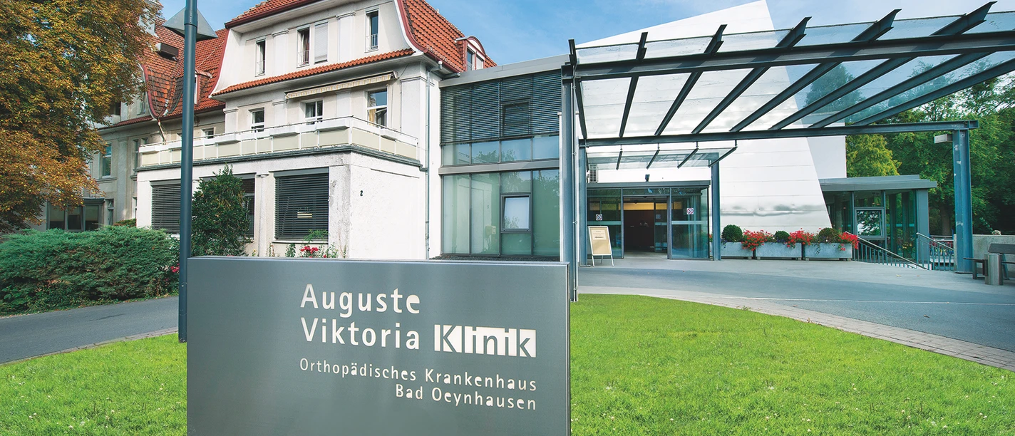 Auguste-Viktoria-Klinik Das Bild zeigt die Auguste-Viktoria-Klinik mit einem modernen Eingangsbereich und einem historischen Gebäude im Hintergrund. Im Vordergrund steht ein Schild mit dem Namen der Klinik, während eine Überdachung den Eingangsbereich überspannt.