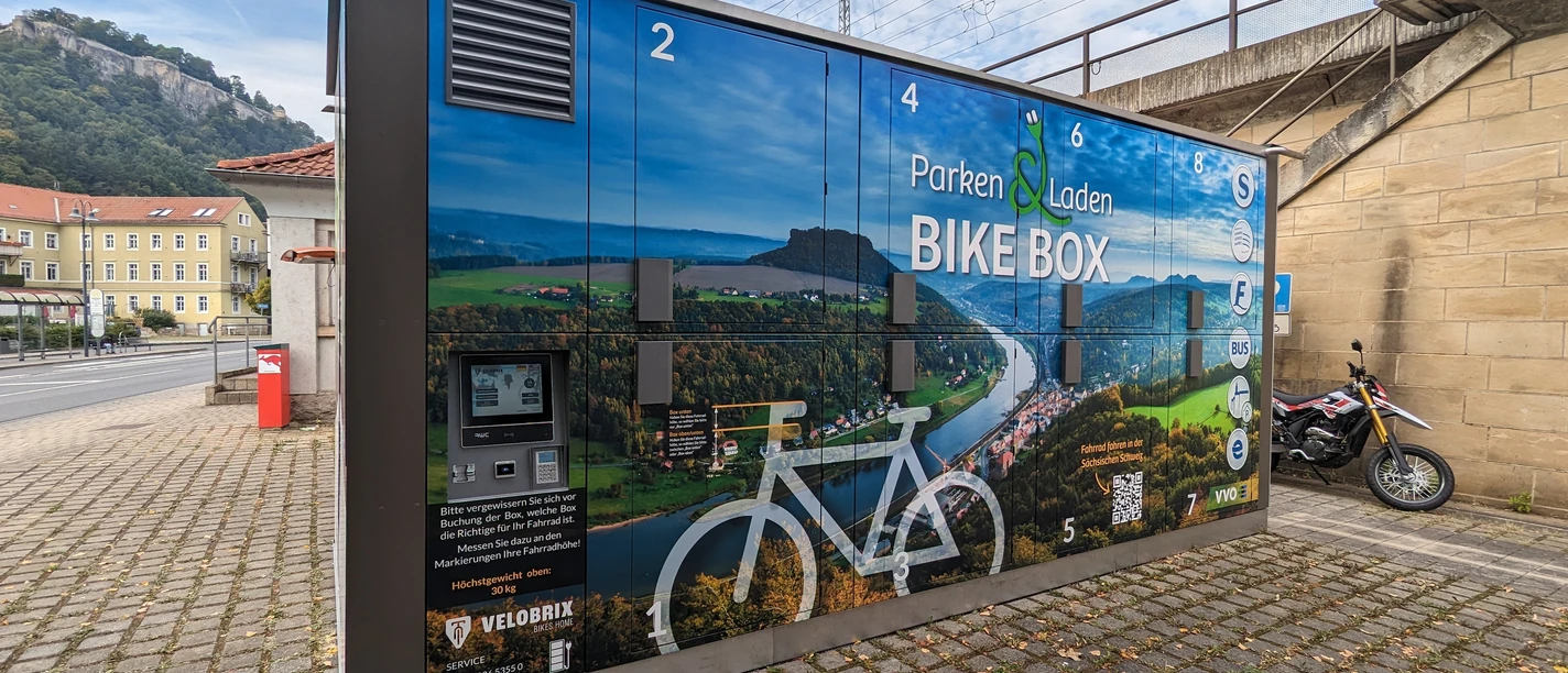 Fahrradgarage Königstein Fahrradbox mit Landschaftsbild und Beschriftung "Parken und Laden" an einer Straße, daneben ein Motorrad, im Hintergrund ein Hügel.