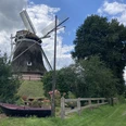 Mühle Burlage, Gemeinde Rhauderfehn, Außenansicht, Ostfriesland Traditionelle Windmühle mit großen Flügeln auf einem Hügel, umgeben von Bäumen, einem Zaun und einem kleinen Boot auf einer Wiese