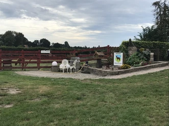 Alpakazucht Büter Eingangsbereich einer Alpakazucht mit rotem Zaun, Hofladen-Schild, Gartenbeet und Sitzgruppe im Grünen.