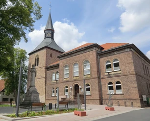 Kirchplatz mit historischer Kirche und Kriegerdenkmal, umgeben von Bäumen unter blauem Himmel.