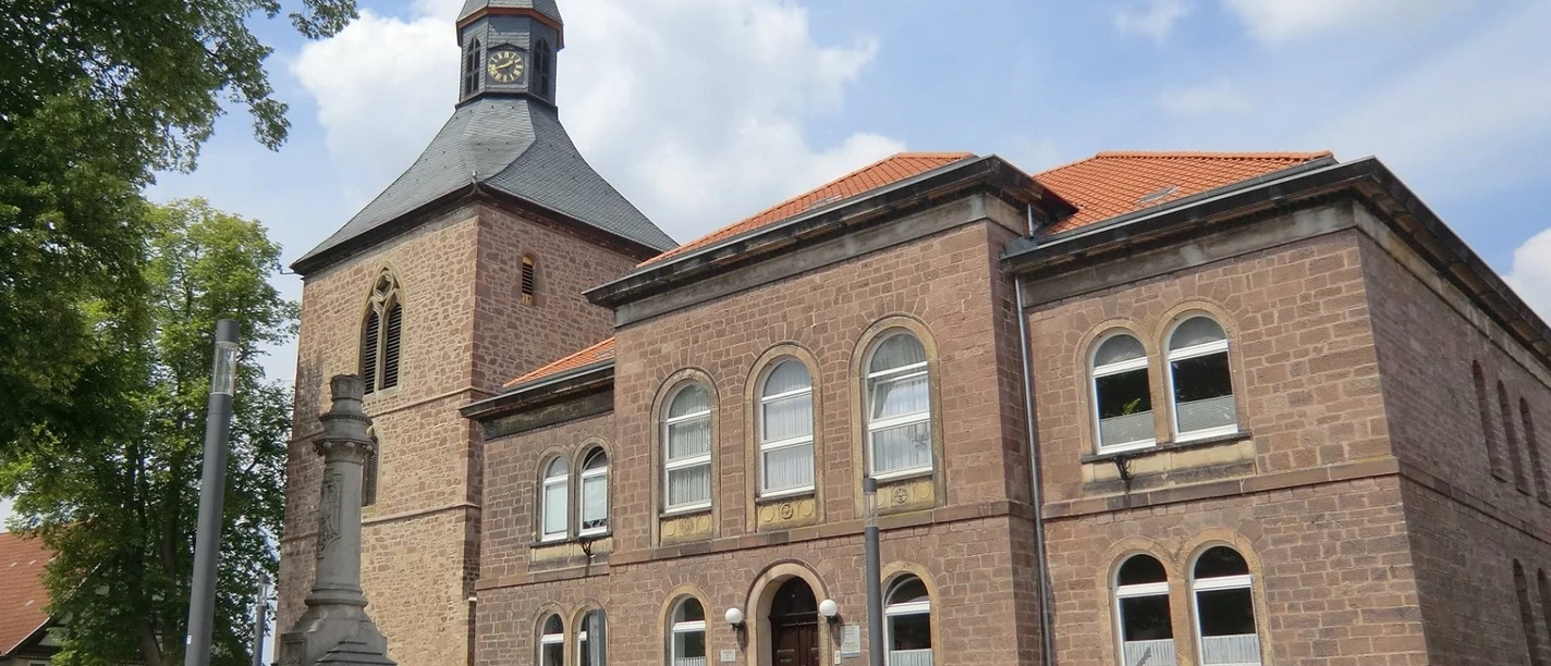 Kirchplatz mit historischer Kirche und Kriegerdenkmal, umgeben von Bäumen unter blauem Himmel.