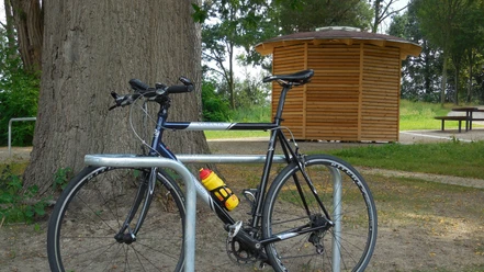 Rad-Rastplatz Retzen Fahrrad steht an Metallständer vor Holzhäuschen, umgeben von Bäumen und einem Picknicktisch.