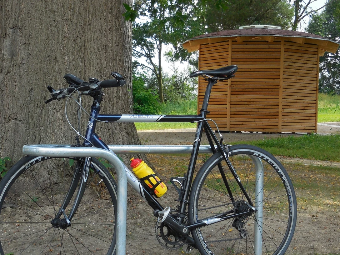 Rad-Rastplatz Retzen Fahrrad steht an Metallständer vor Holzhäuschen, umgeben von Bäumen und einem Picknicktisch.