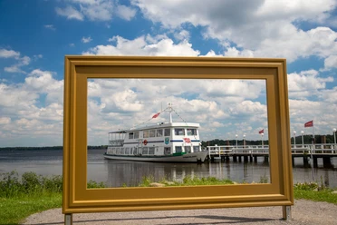 Das Bild zeigt die Weiße Flotte, aus der Sicht durch einen goldenen Bilderrahmen. Im Hintergrund erstreckt sich ein blauer Himmel mit vereinzelten Wolken. Das Schiff liegt an einem Steg vor Anker, umgeben von ruhigem Wasser und grüner Ufervegetation.