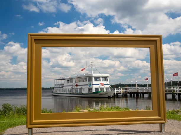 Das Bild zeigt die Weiße Flotte, aus der Sicht durch einen goldenen Bilderrahmen. Im Hintergrund erstreckt sich ein blauer Himmel mit vereinzelten Wolken. Das Schiff liegt an einem Steg vor Anker, umgeben von ruhigem Wasser und grüner Ufervegetation.