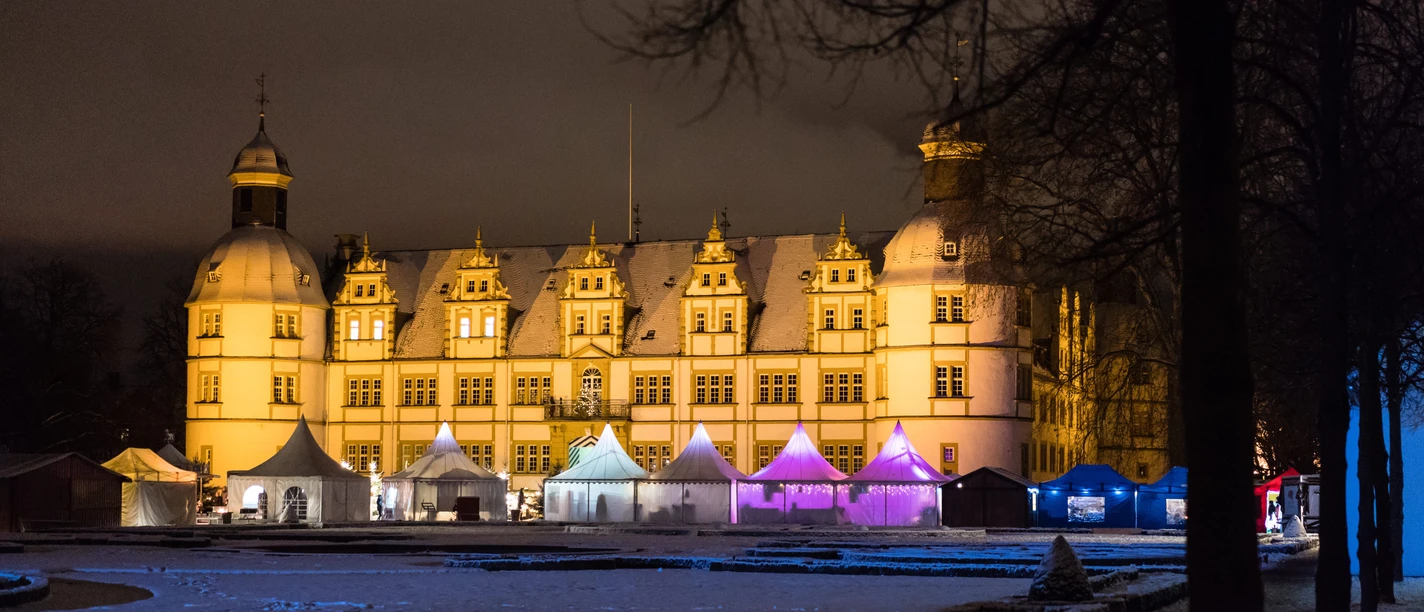 Beleuchtetes Schloss im Winter, umgeben von festlich beleuchteten Marktständen im Hintergrund.