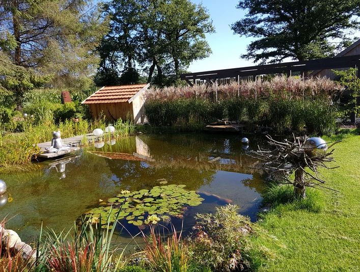 20190922_110739.jpg Ein ruhiger Teich spiegelt inmitten eines gepflegten Gartens lilienbedeckte Wasserflächen und harmonische Vegetation wider, während eine Statue am Ufer sitzt und für Harmonie sorgt.