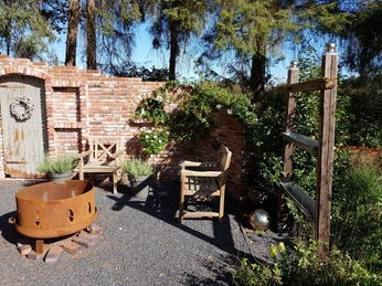 20190922_111109.jpg Auf dem Bild ist ein rustikaler Gartenbereich zu sehen. Eine Backsteinmauer mit dekorativen Elementen bietet einen charmanten Hintergrund. Zwei Holzgartenstühle laden zum Verweilen ein und ein stilvoller Feuerkorb sorgt für gemütliche Atmosphäre. Umliegende Pflanzen verstärken das naturnahe Ambiente.
