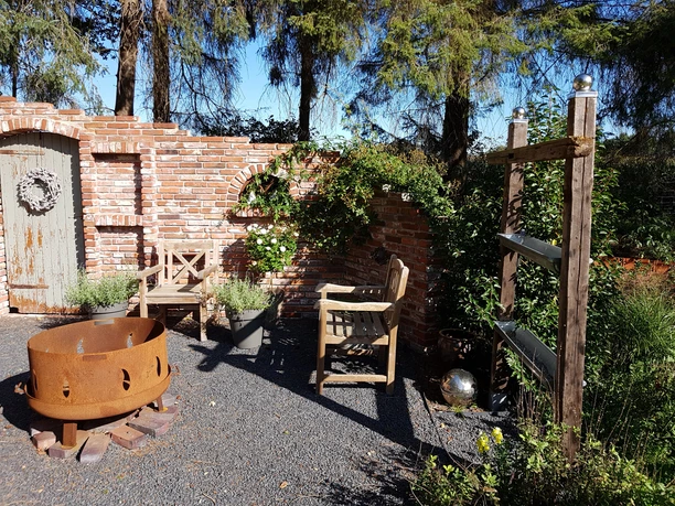 20190922_111109.jpg Auf dem Bild ist ein rustikaler Gartenbereich zu sehen. Eine Backsteinmauer mit dekorativen Elementen bietet einen charmanten Hintergrund. Zwei Holzgartenstühle laden zum Verweilen ein und ein stilvoller Feuerkorb sorgt für gemütliche Atmosphäre. Umliegende Pflanzen verstärken das naturnahe Ambiente.