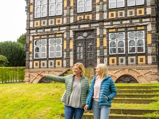 Zwei Frauen in farbigen Jacken spazieren vor einem historischen Fachwerkhaus mit dekorativen Fenstern.