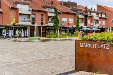 Marktplatz mit Schriftzug Roter Ziegelmarktplatz mit modernen Gebäuden, Radfahrern und Wasserspielen in einer lebhaften Stadt.
