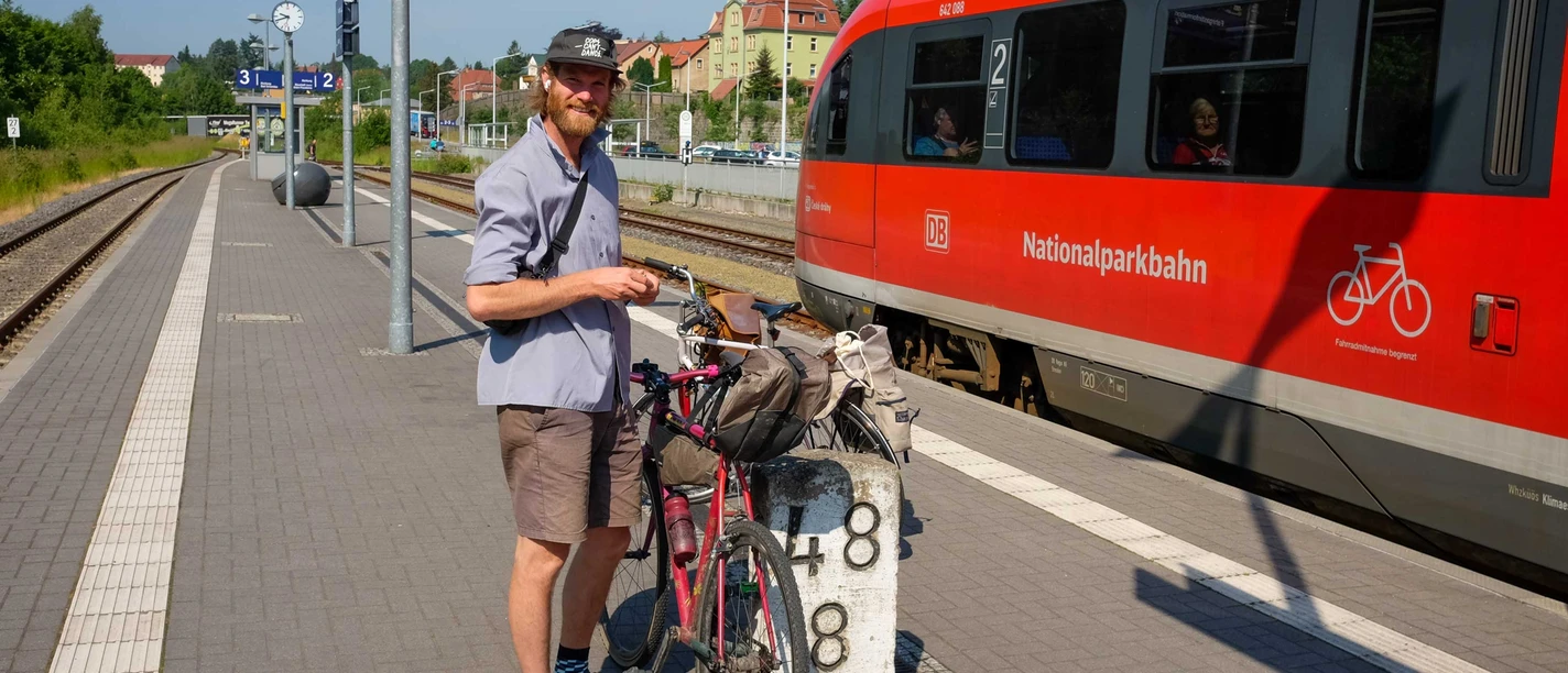 Bahnhof Sebnitz mit dem Fahrrad Mann mit Fahrrad steht an einem Bahnsteig, roter Zug fährt ein.
