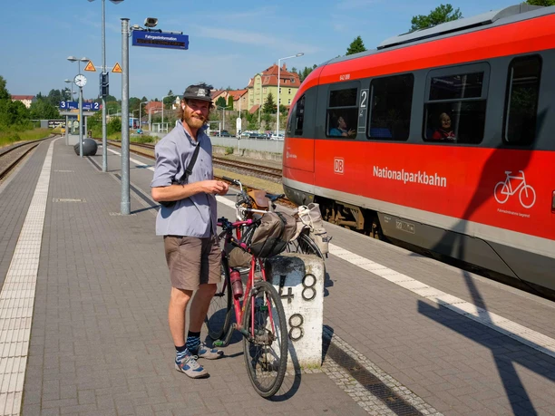 Bahnhof Sebnitz mit dem Fahrrad Mann mit Fahrrad steht an einem Bahnsteig, roter Zug fährt ein.