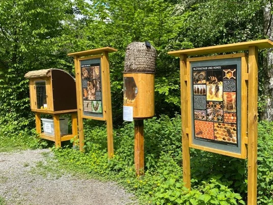 Bienenlehrpfad in Erkrath Drei hölzerne Informationstafeln und Bienenbehausungen stehen inmitten einer grünen Landschaft.