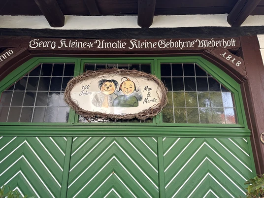 Grüne Holz-Eingangstür des Wilhelm Busch Museums mit einem Schild von Max und Moritz, umrahmt von Ästen.