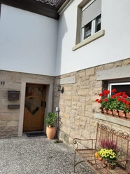 Eingangsbereich einer Ferienwohnung mit Holztür, blühenden Geranien und einer schmiedeeisernen Bank.