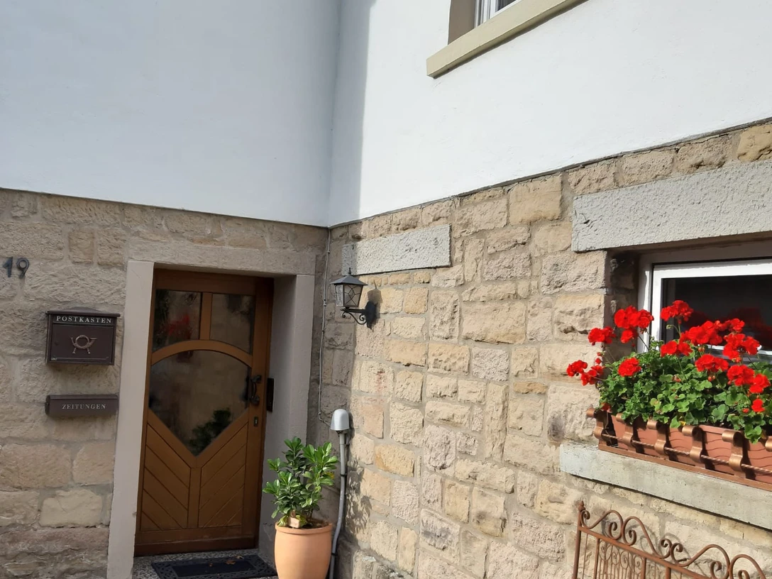 Eingangsbereich einer Ferienwohnung mit Holztür, blühenden Geranien und einer schmiedeeisernen Bank.