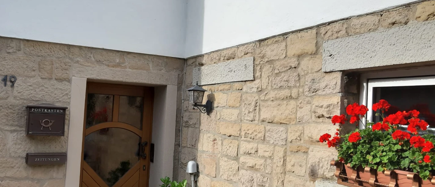 Ferienwohnung_AmNelkenweg.jpg Eingangsbereich einer Ferienwohnung mit Holztür, blühenden Geranien und einer schmiedeeisernen Bank.