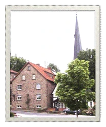 Ferienwohnung Capella.gif Ein Backsteinhaus mit grünem Baum im Vordergrund, dahinter ein Kirchturm in ländlicher Umgebung.