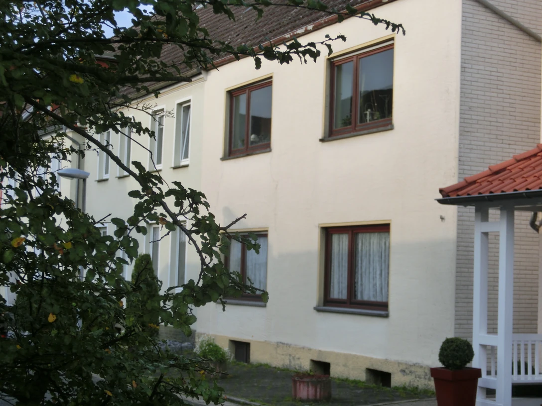 Mehrfamilienhaus mit verputzter Fassade, mehreren Stockwerken und einfachen Fenstern, umgeben von Bäumen.