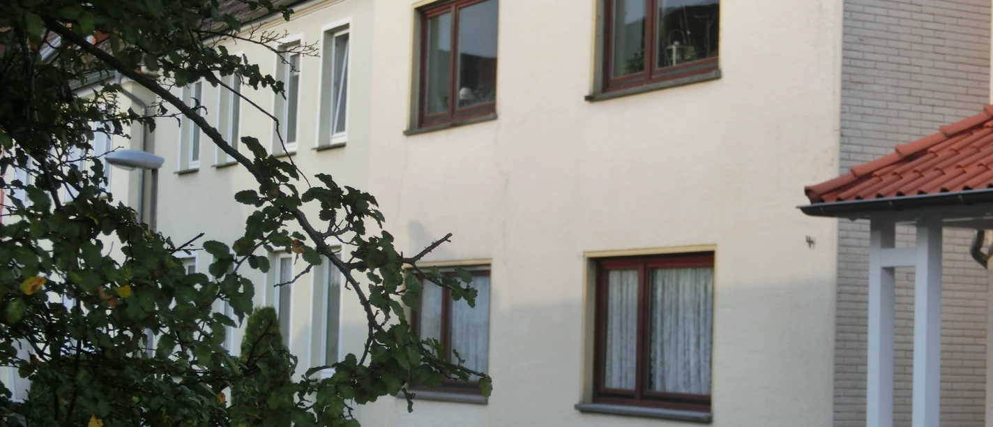 Mehrfamilienhaus mit verputzter Fassade, mehreren Stockwerken und einfachen Fenstern, umgeben von Bäumen.