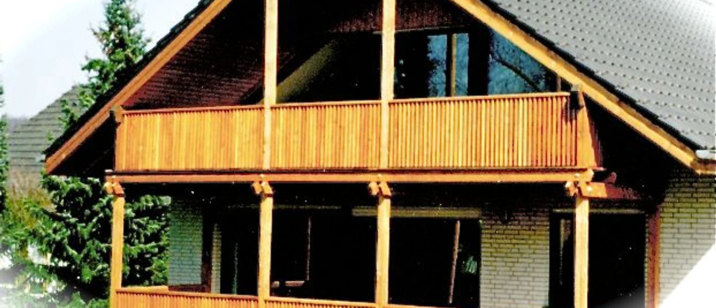 Zweistöckiges Haus mit großen Holzbalkonen und schrägem Dach, umgeben von Nadelbäumen.