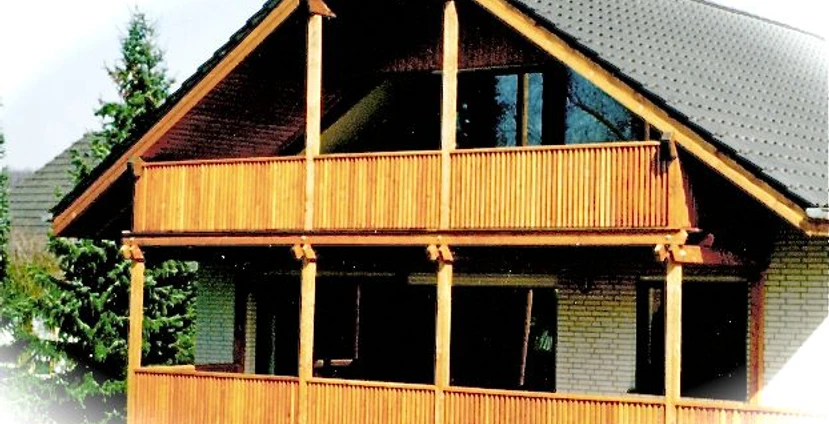 Zweistöckiges Haus mit großen Holzbalkonen und schrägem Dach, umgeben von Nadelbäumen.
