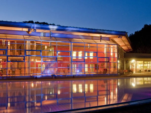 Toskana Therme Bad Schandau_Frank Höhler.jpg Die Toskana Therme in Bad Schandau zeigt ein beleuchtetes Gebäude mit einer gläsernen Fassade im Abendlicht, umgeben von einer reflektierenden Wasseroberfläche.