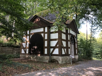 Fachwerkpavillon im Grünen mit steinerner Basis und zwei großen, dunkelbraunen Holztüren.