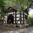 Fachwerkpavillon im Grünen mit steinerner Basis und zwei großen, dunkelbraunen Holztüren.