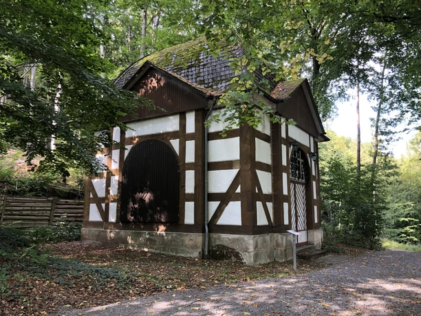 Friedrichspavillon.jpg Fachwerkpavillon im Grünen mit steinerner Basis und zwei großen, dunkelbraunen Holztüren.