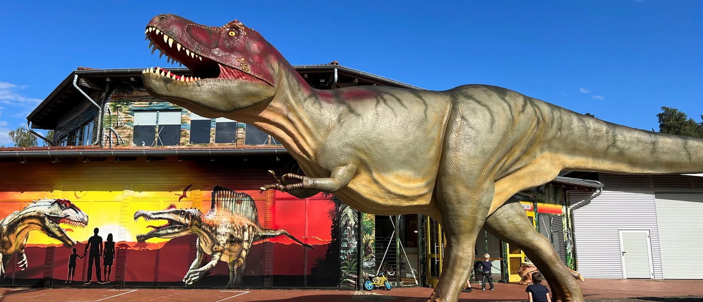 Dinopark-Münchehagen.jpg En dinosaurmodel i naturlig størrelse står foran en rød bygning i Dinopark Münchehagen.