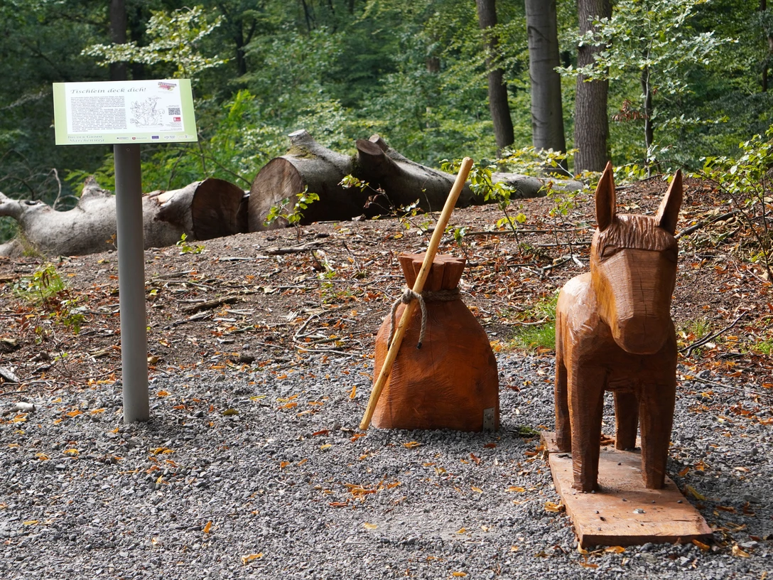 Holzfiguren von Esel und Beutel des Märchens Tischlein deck dich im bewaldeten Bereich am Wegesrand.