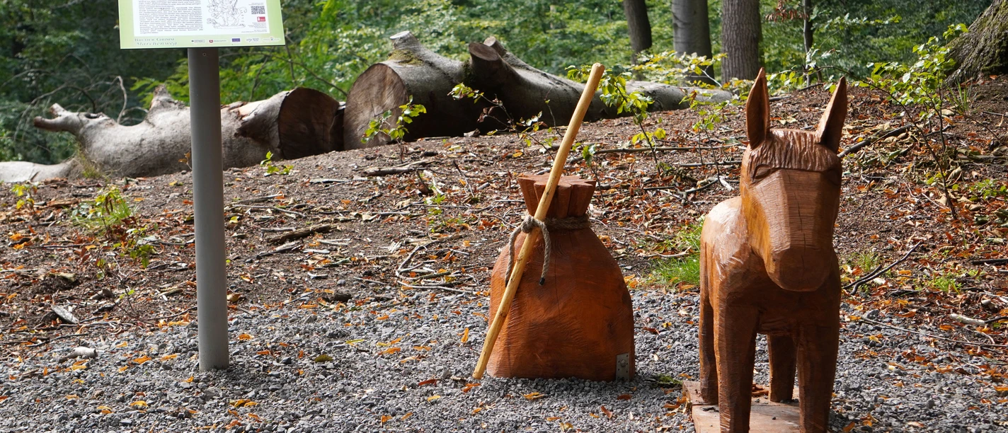 Tischlein-Deck-Dich-Märchenfigur.jpg Holzfiguren von Esel und Beutel des Märchens Tischlein deck dich im bewaldeten Bereich am Wegesrand.