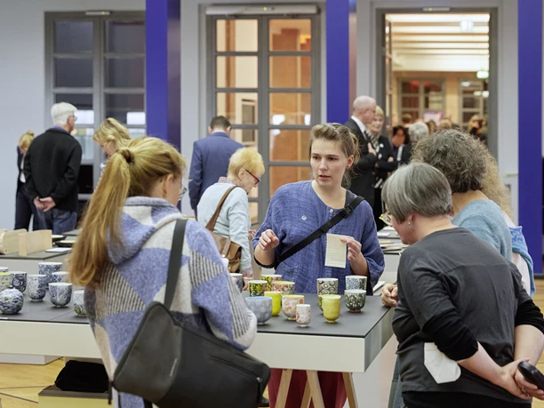 Personen betrachten handgefertigte Keramik auf einer Kunstmesse in einem hellen Ausstellungsraum.