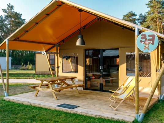 Safari-Zelt-Lodge Ein Safari-Zelt mit Holzterrasse, Sitzbank und zwei Liegestühlen auf grünem Wiesengelände.