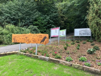 Holzschild mit der Aufschrift "Brüder Grimm Wanderweg" am Eingang zu einem mit Tafeln gesäumten Pfad.
