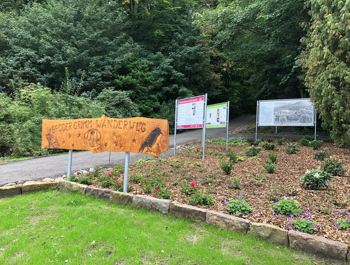 Start Märchenweg Holzschild mit der Aufschrift "Brüder Grimm Wanderweg" am Eingang zu einem mit Tafeln gesäumten Pfad.