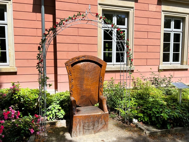 Ein uriger Holzthron von einem Rosenbogen umgeben, auf einem gepflasterten Weg vor einem rosa Gebäude.