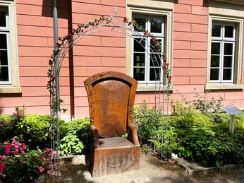 Märchenthron.jpg Ein uriger Holzthron von einem Rosenbogen umgeben, auf einem gepflasterten Weg vor einem rosa Gebäude.