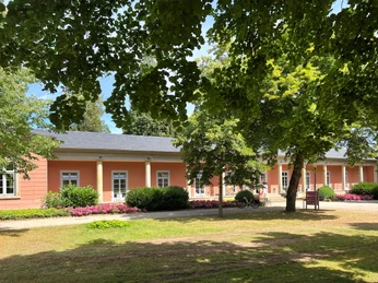 Romantik-Bad-Rehburg-(1).jpg Ein historisches, rosa gefärbtes Kurhaus mit hohen Fenstern und blühenden Blumen in sonniger Umgebung.