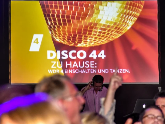 WDR 4 - Disco 44_mit Logo.jpg DJ steht vor einer großen Leinwand, auf der "Disco 44" und das WDR 4-Logo projiziert sind.