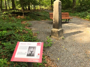 Holzbank und historischer Stein an einem Schotterpfad im Wald. Informationstafel im Vordergrund.