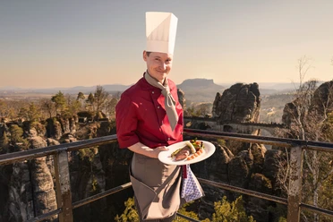 A chef in a red uniform and white cap presents a dish on a plate, with a rocky landscape at sunset in the background.