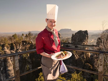 Koch Panoramarestaurant Bastei Ein Koch in roter Uniform und weißer Mütze präsentiert ein Gericht auf einem Teller, im Hintergrund eine felsige Landschaft bei Sonnenuntergang.A chef in a red uniform and white cap presents a dish on a plate, with a rocky landscape at sunset in the background.Kuchař v červené uniformě a bílé čepici předkládá pokrm na talíři, v pozadí je skalnatá krajina při západu slunce.Szef kuchni w czerwonym mundurze i białej czapce prezentuje danie na talerzu, a w tle widać skalisty krajobraz o zachodzie słońca.Een chef-kok in een rood uniform en witte pet presenteert een gerecht op een bord, met op de achtergrond een rotslandschap bij zonsondergang.Uno chef in uniforme rossa e berretto bianco presenta un piatto su un piatto, con un paesaggio roccioso al tramonto sullo sfondo.