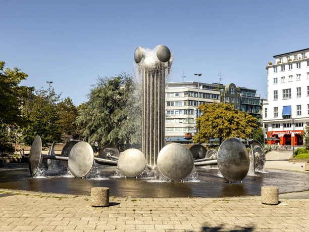 Brunnen am Ebertplatz Der Ebertplatz in Köln zeigt einen modernen Brunnen mit Wassertrommeln und einer zentralen Säule. Umgeben von Bäumen und Gebäuden, bietet er eine urbane Kulisse an einem sonnigen Tag.