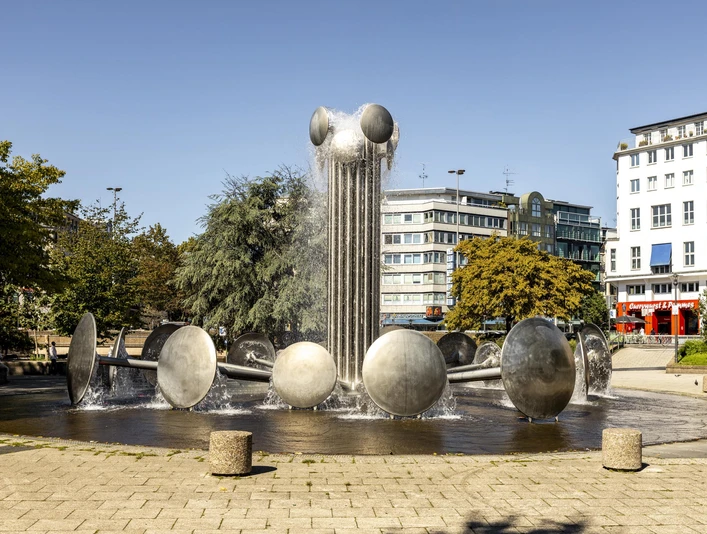 Ebertplatz Der Ebertplatz in Köln zeigt einen modernen Brunnen mit Wassertrommeln und einer zentralen Säule. Umgeben von Bäumen und Gebäuden, bietet er eine urbane Kulisse an einem sonnigen Tag. Ebertplatz in Cologne features a modern fountain with water drums and a central column. Surrounded by trees and buildings, it provides an urban backdrop on a sunny day.
